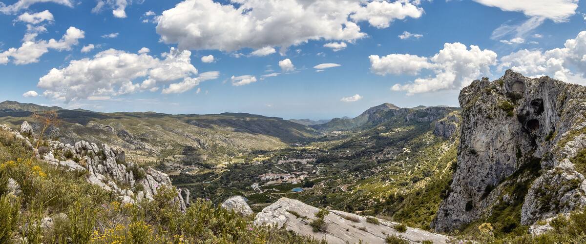 A mountainous landscape with a rocky outcrop, a valley with scattered buildings, and hikers on a trail.