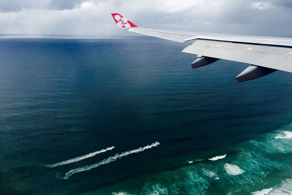A asa de um avião com o logotipo da Air Asia, sobrevoando um vasto oceano com turbulência visível na esteira.