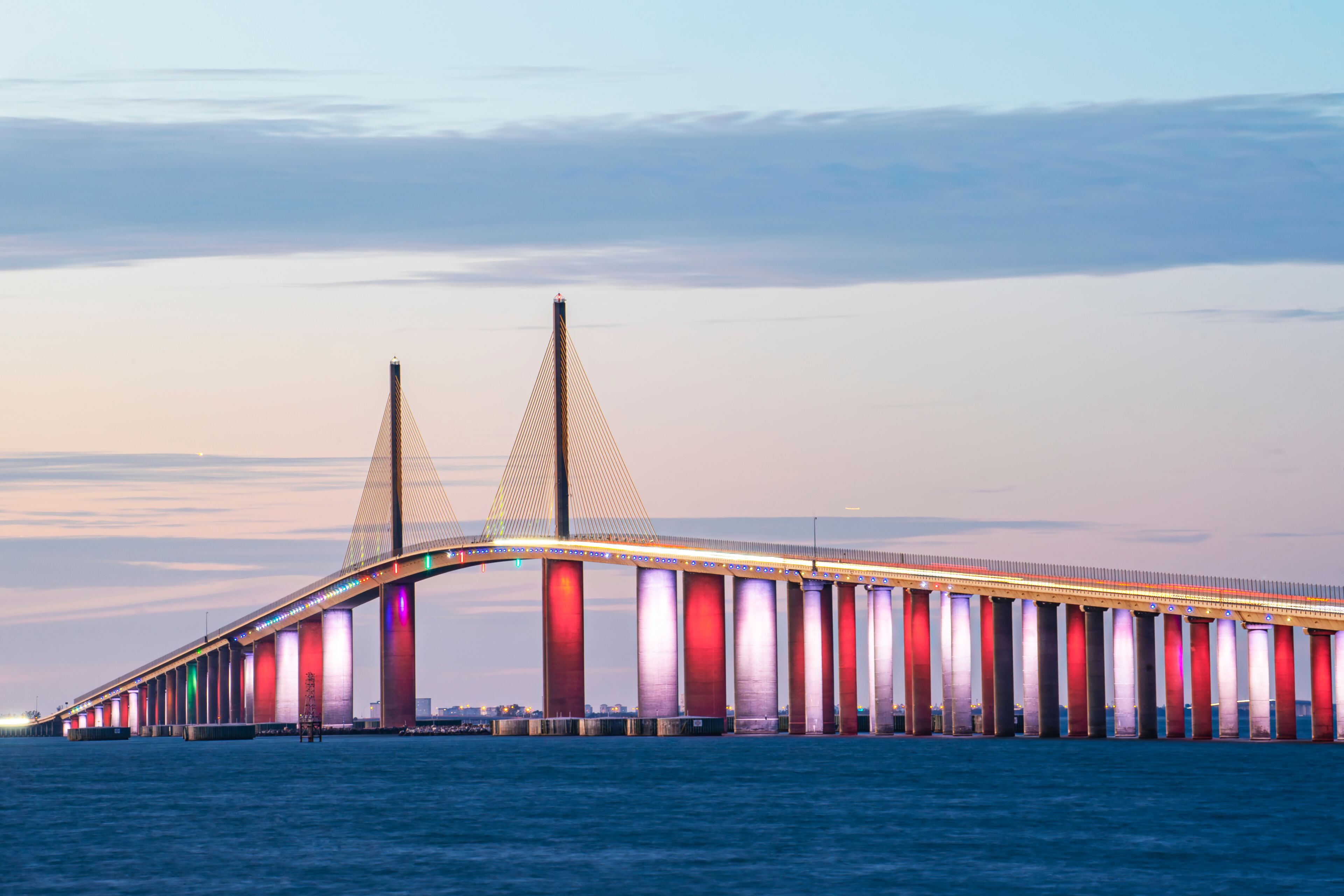 Sunshine Skyway Bridge over Tampa Bay in Florida, USA, at sunset. Red, White, and Blue lights shine under the bridge, and a pastel sky is visible behind the bridge. The water is dark navy below