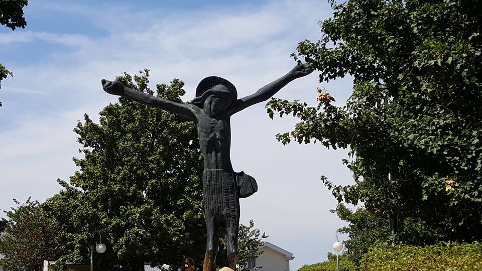 A crucifixion statue with outstretched arms, surrounded by trees and a clear sky.