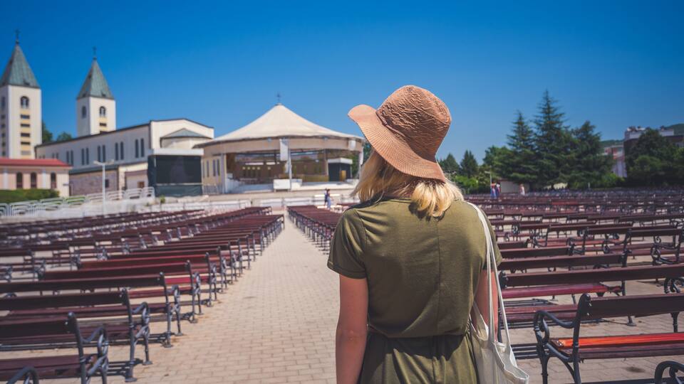 A woman in a hat walking down a row of empty benches towards a church with two towers.