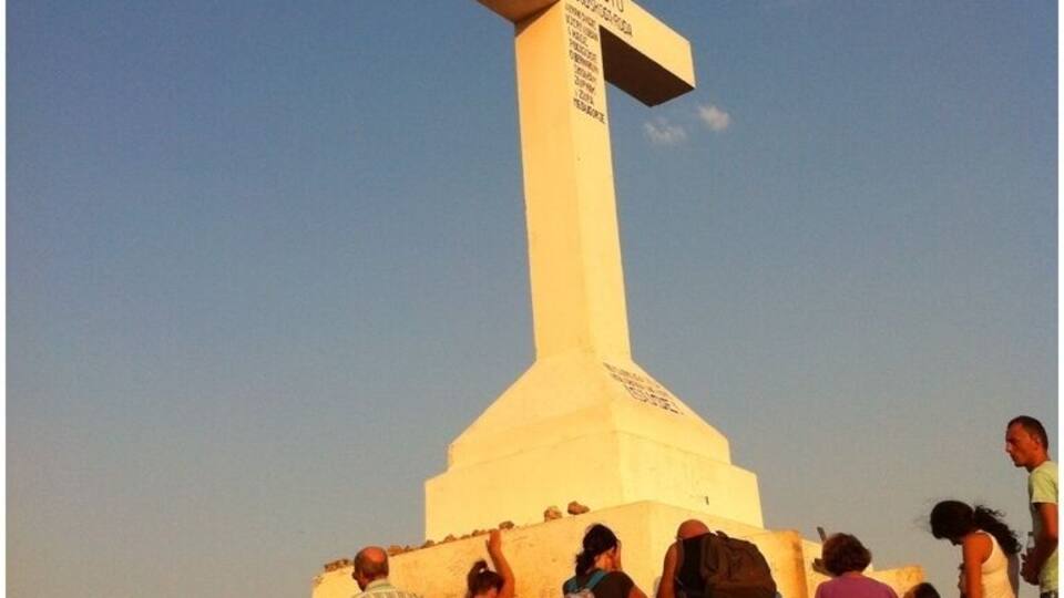 A large white cross with inscriptions, surrounded by a crowd of people.