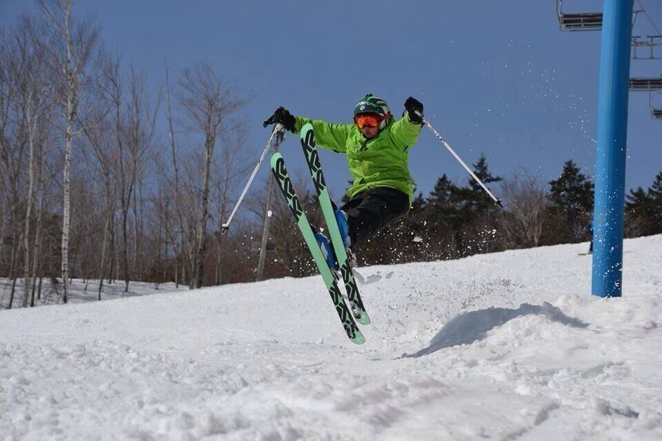 A skier in mid-air performing a jump with skis and poles, surrounded by snow and trees.