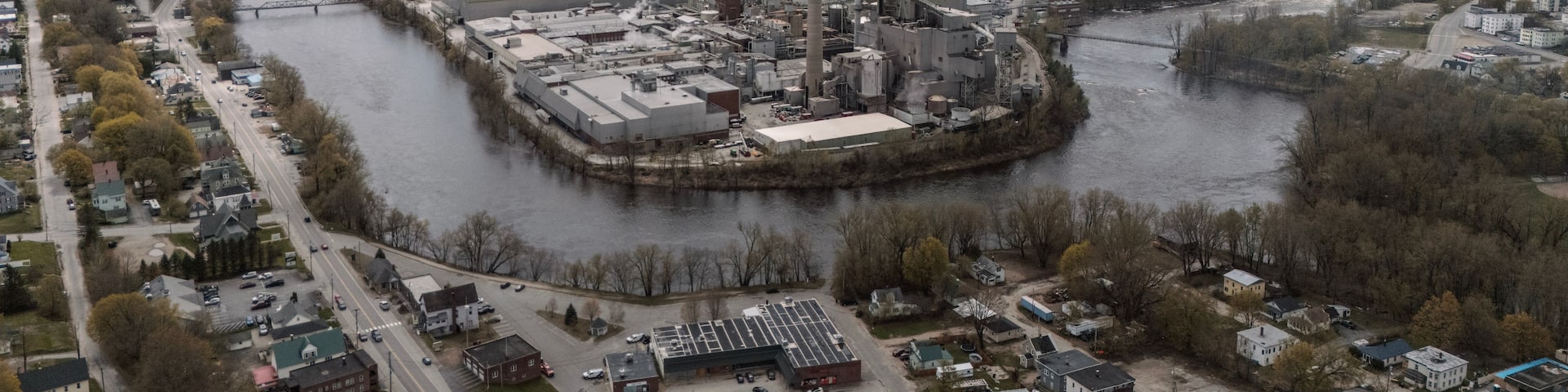 An industrial complex by a river with multiple smokestacks, surrounded by a town and mountains in the background.