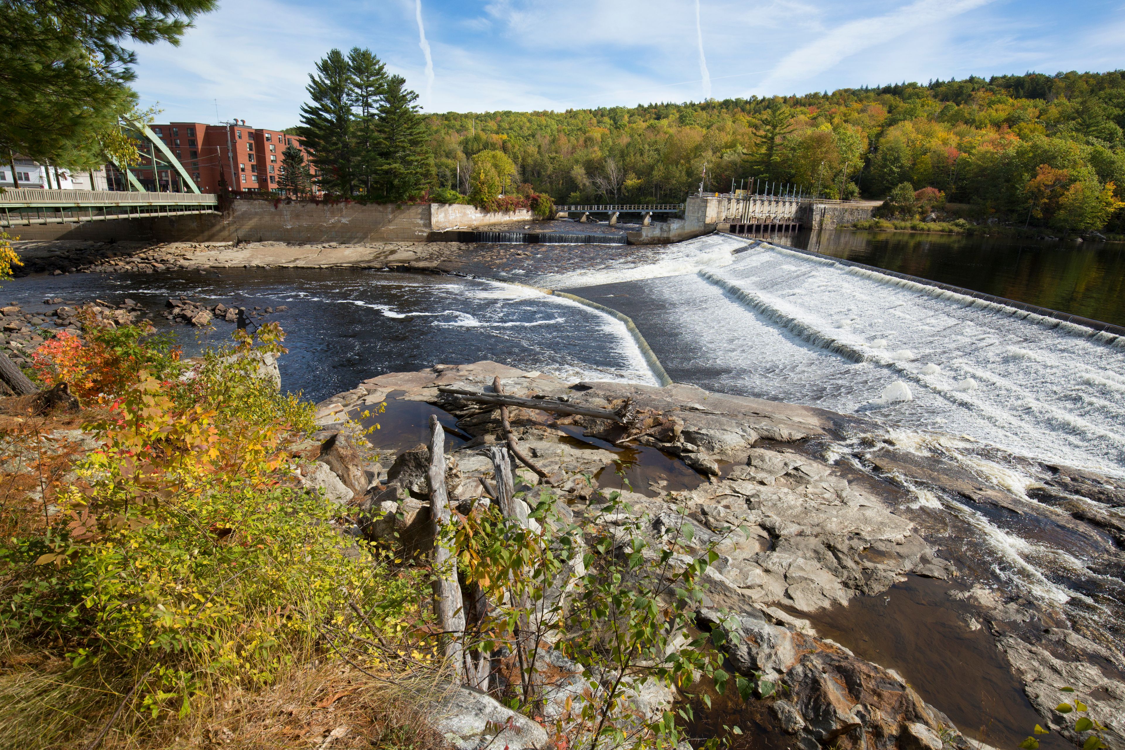 A dam with water flowing over it, surrounded by autumn foliage and a bridge.
