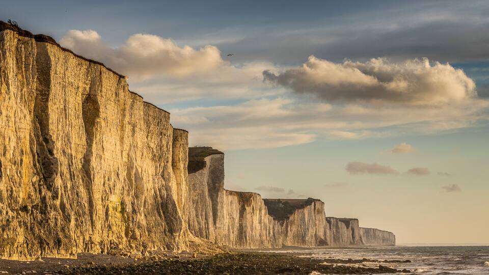 A coastal cliff with a layered structure, a visible sea, and a partly cloudy sky.