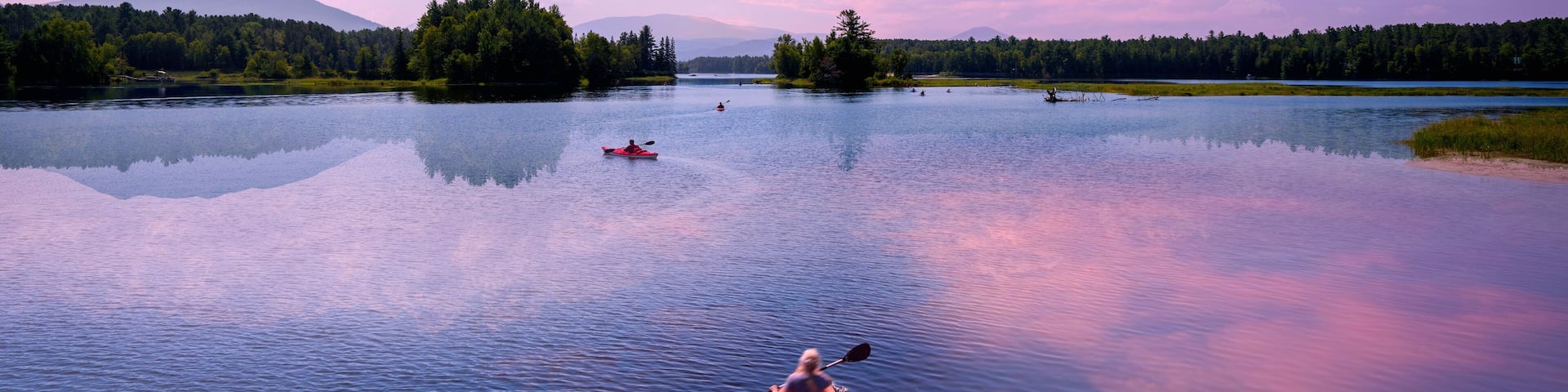A calm lake with kayakers, surrounded by forested hills and a clear sky.