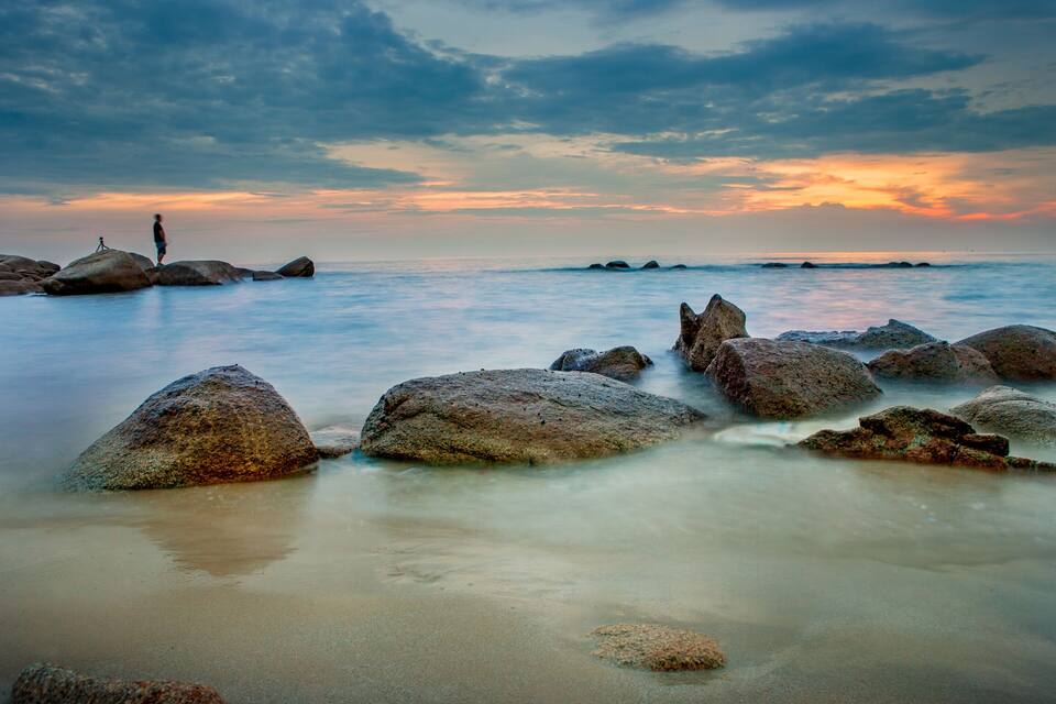 A beach with large rocks and a person standing on one of them.