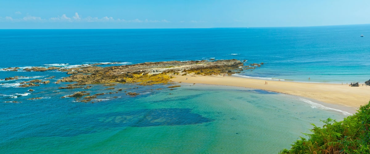 A coastal landscape with clear turquoise waters, a sandy beach, and rocky outcrops.