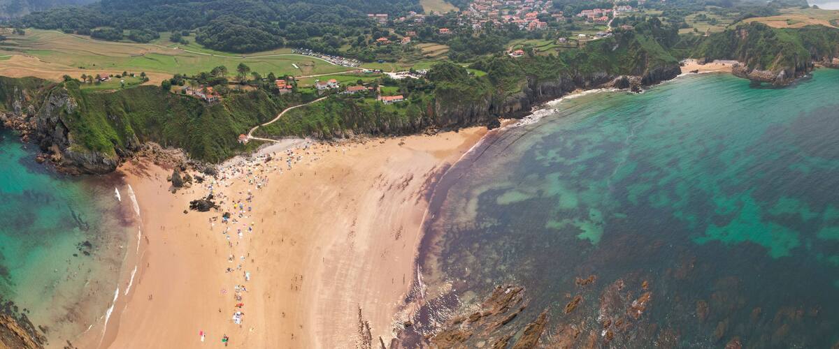 An aerial view of a coastal area with a sandy beach, rocky cliffs, and a small town with houses and greenery.