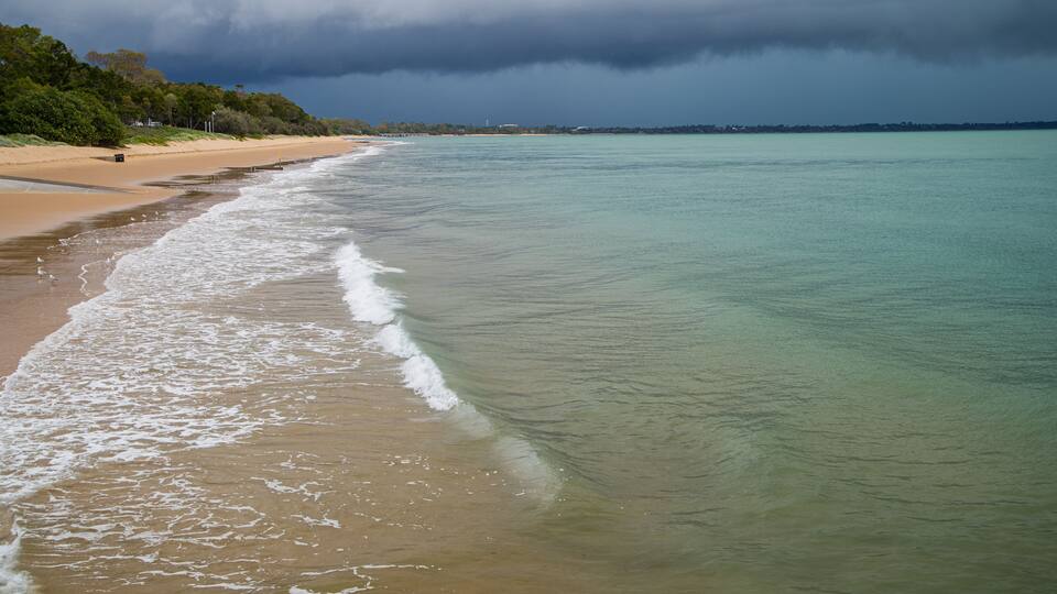 Une plage à l’eau turquoise et limpide, bordée d’un rivage sablonneux, avec une zone boisée en arrière-plan.