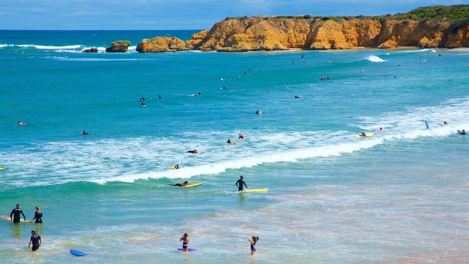A crowded beach with surfers and swimmers in the water, and a rocky cliff in the background.