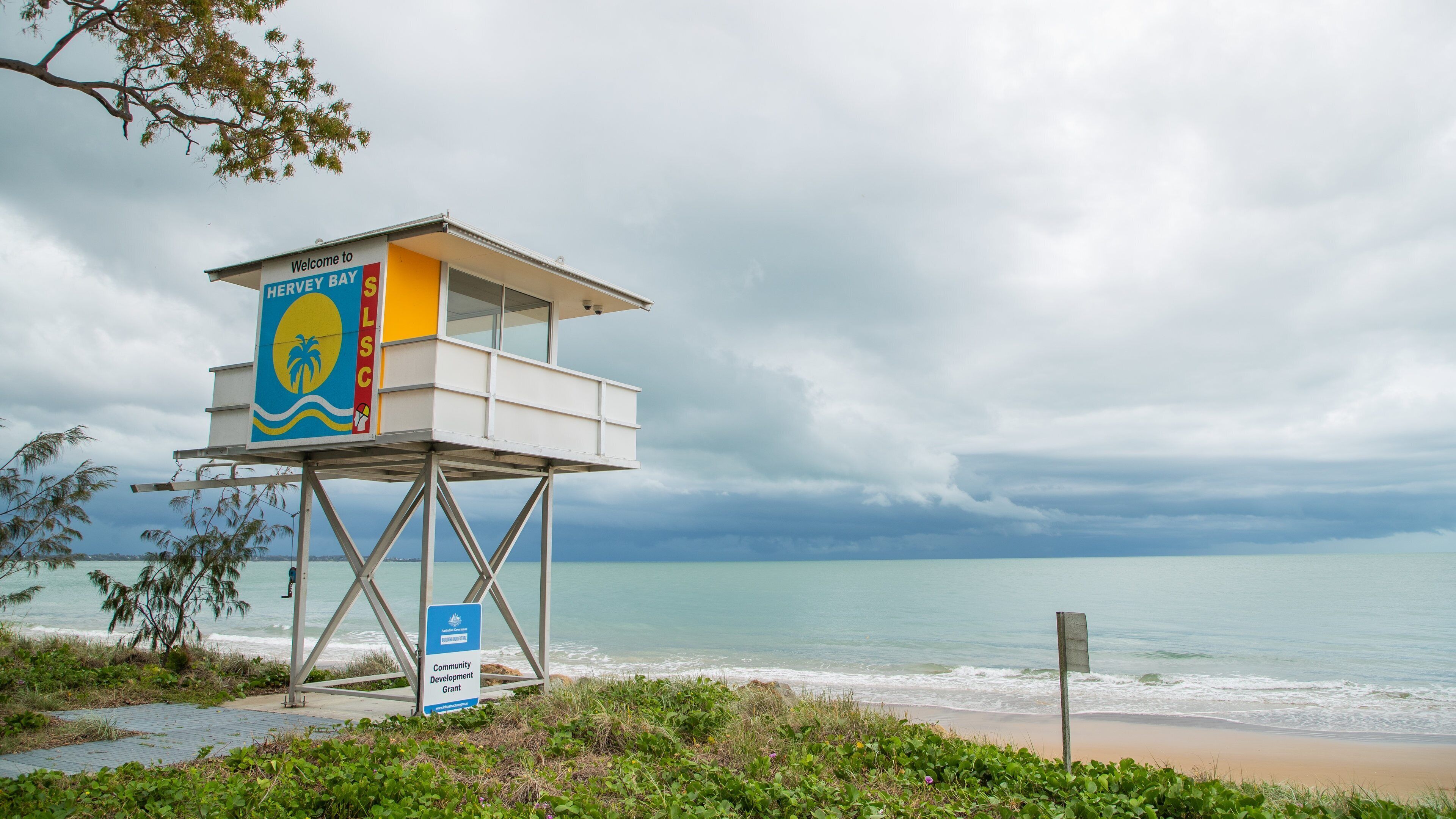 A lifeguard tower with a 'Welcome to HERRY BAY' sign, a beach, and the ocean under cloudy skies.