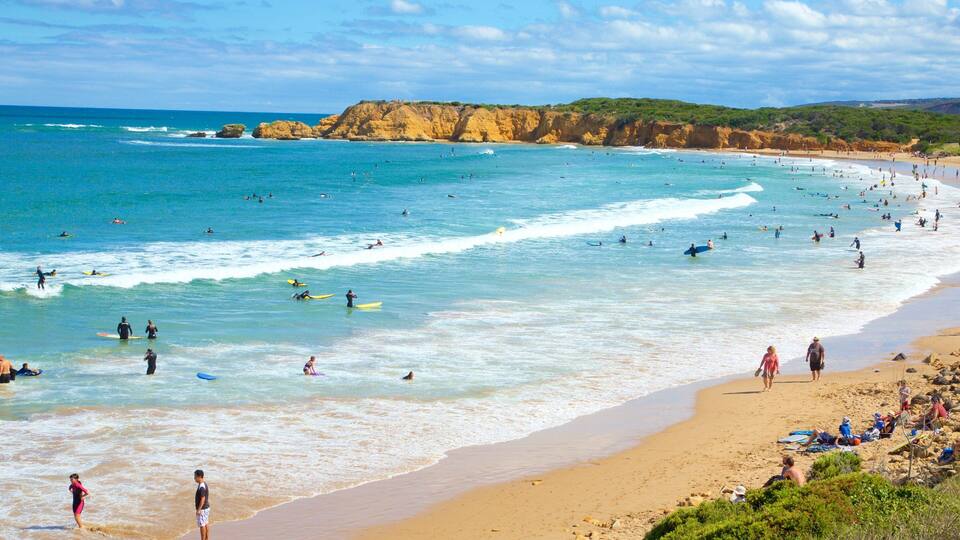 A crowded beach with surfers and swimmers in the water, a sandy shore, and cliffs in the background.