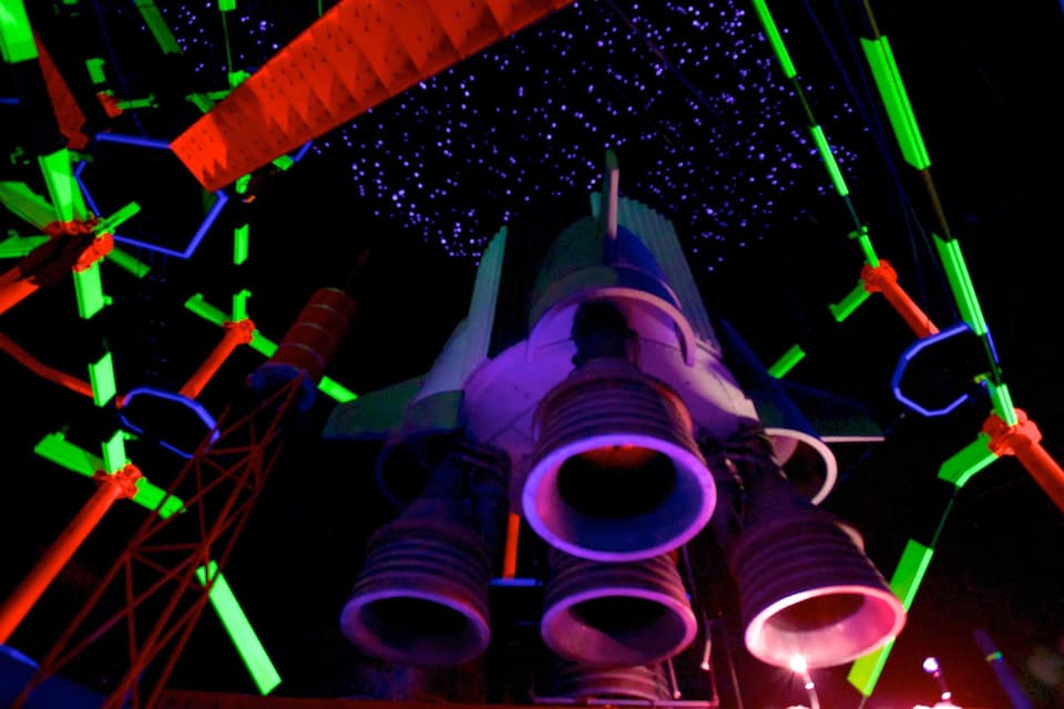 A space shuttle with multiple rocket engines, surrounded by colorful lights and structures.
