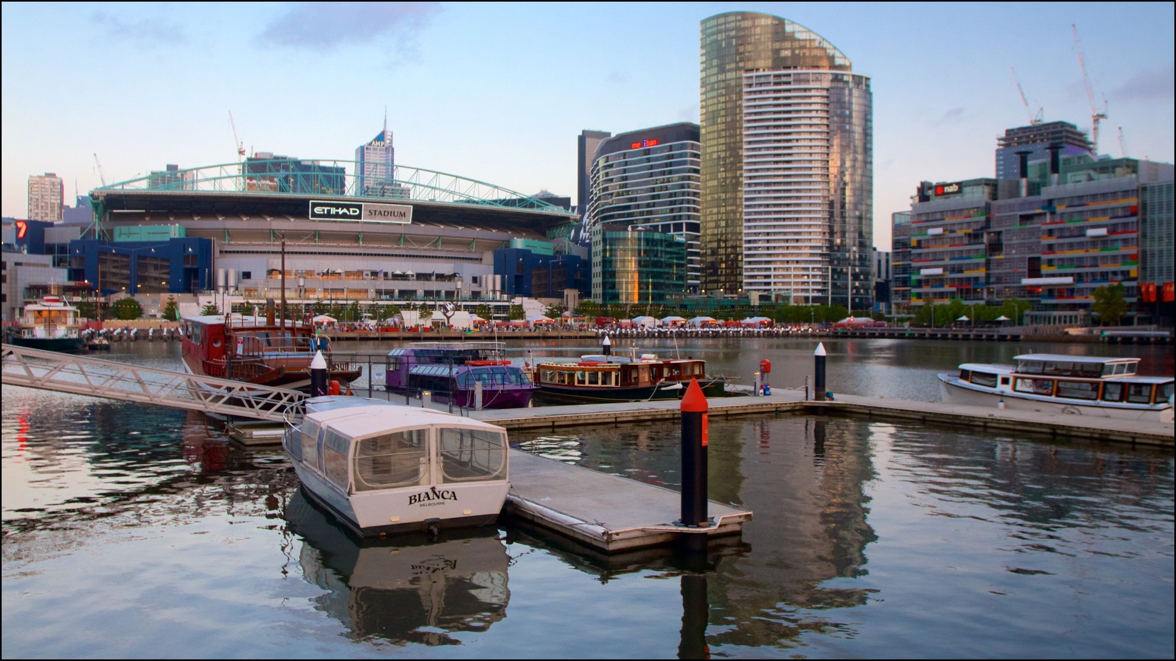 A waterfront with boats docked, a stadium, and modern buildings in the background.