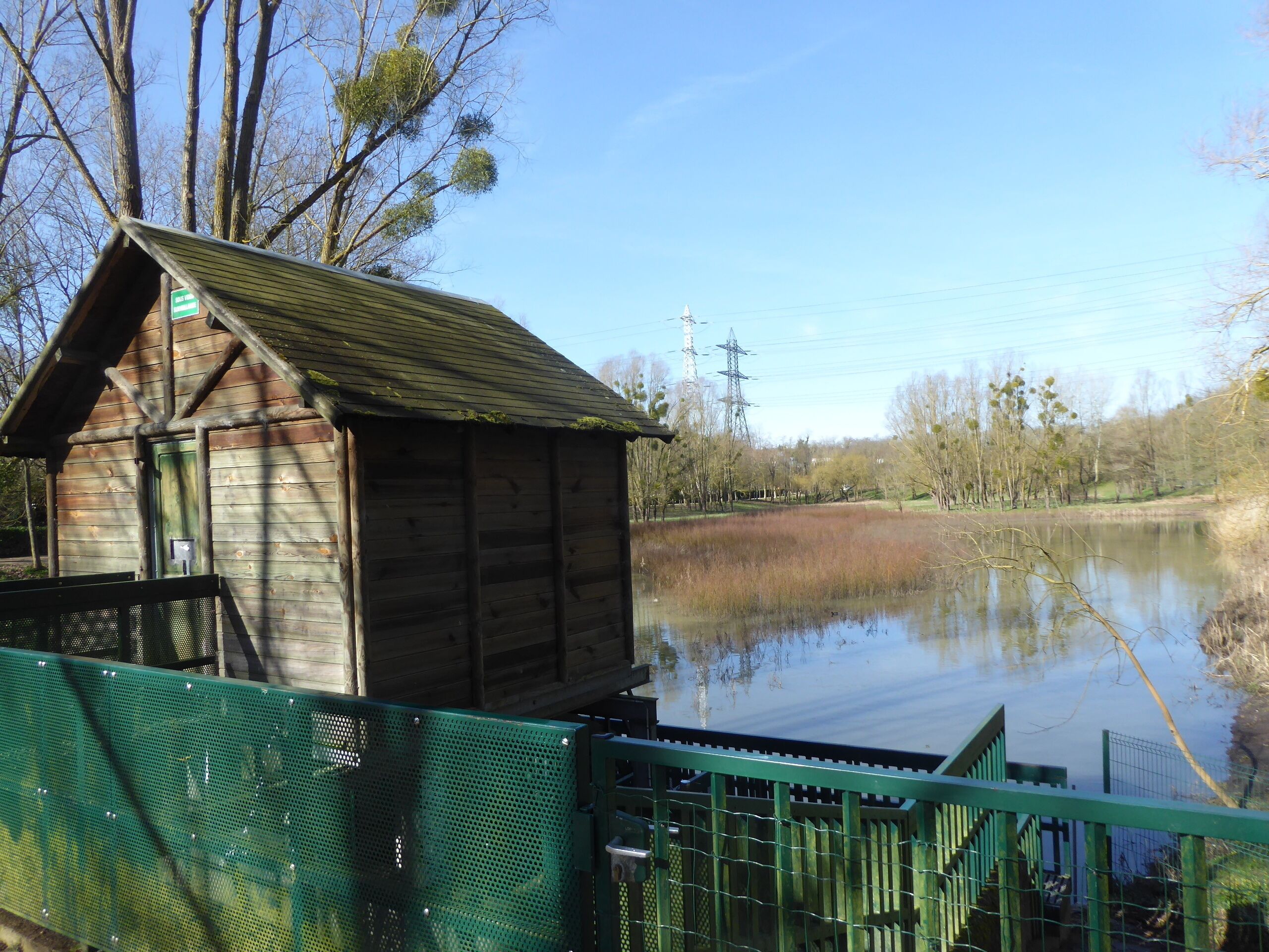 A wooden boathouse by a calm lake with a green metal fence.