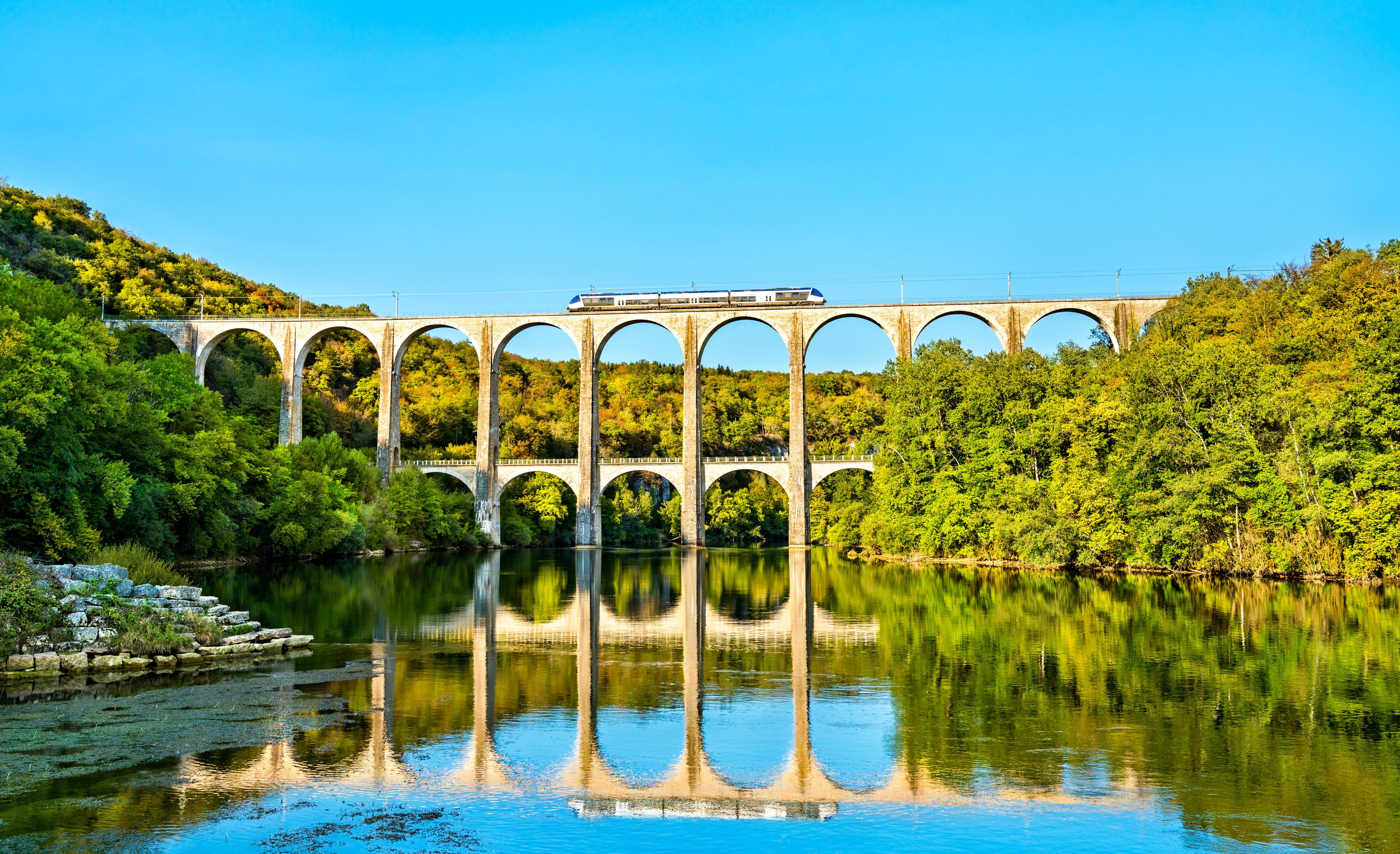 Un pont de pierre enjambant une rivière, avec un train qui le traverse, le tout entouré d’une végétation luxuriante.