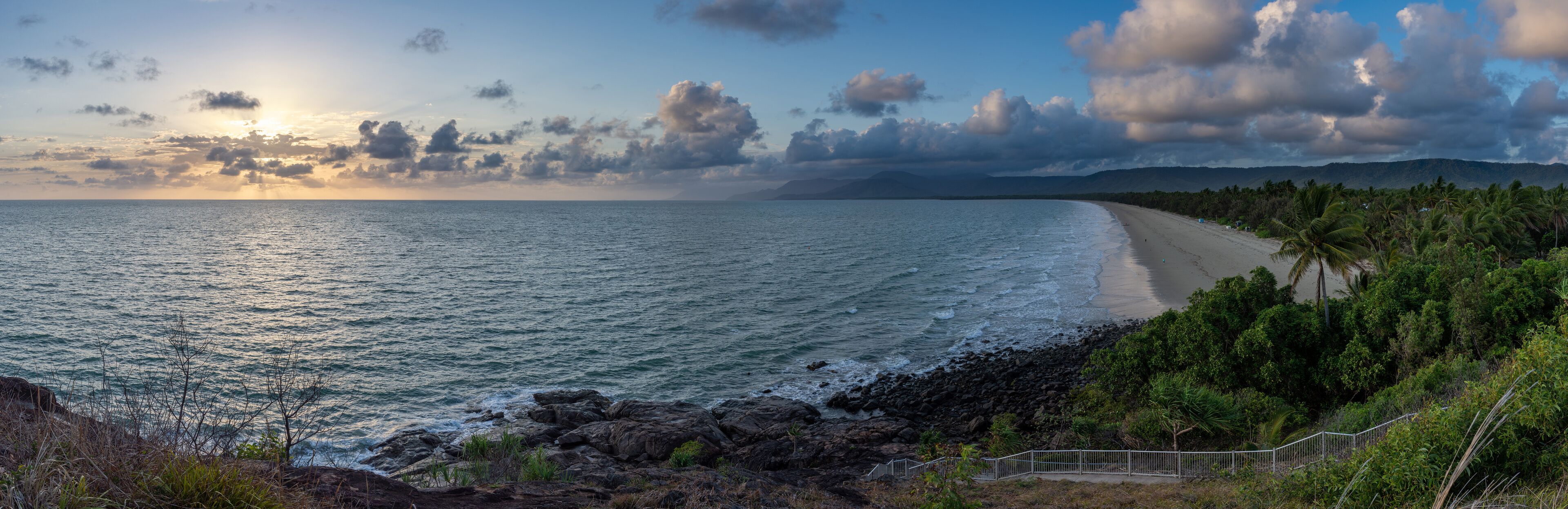 A coastal landscape with a beach, rocky shoreline, and palm trees.