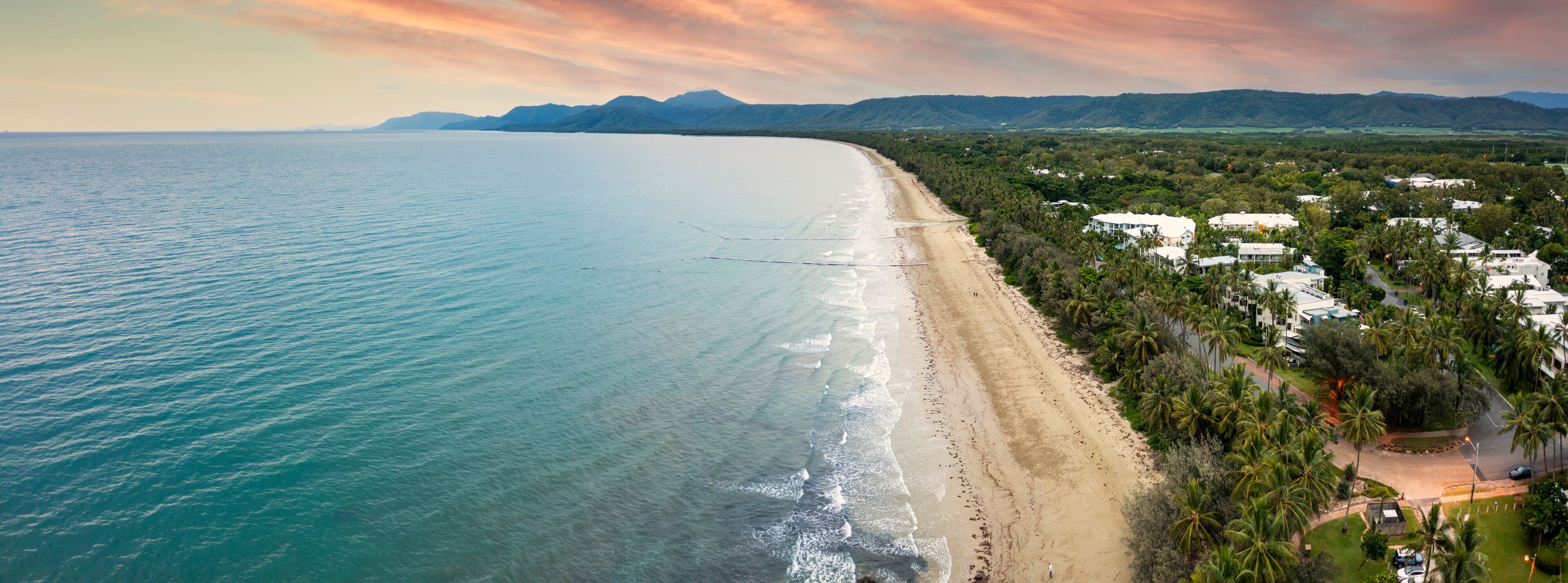 A coastal landscape with a beach, ocean, and a residential area with houses and palm trees.
