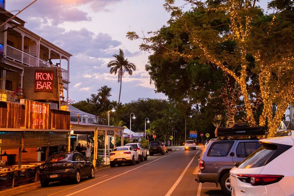 A street lined with trees decorated with lights, a bar named 'IRON BAR', and parked cars.
