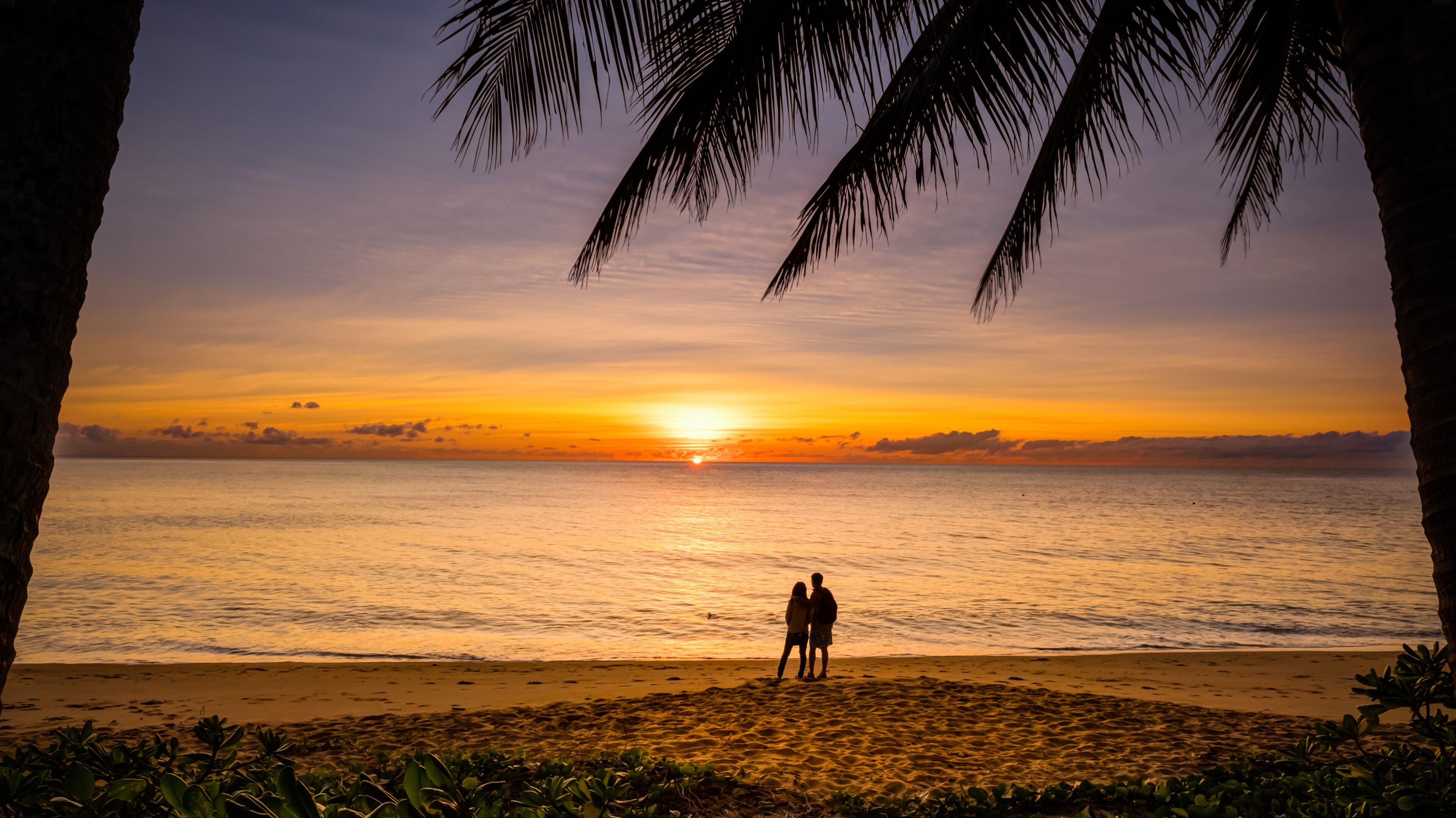 Un atardecer sobre un mar tranquilo con dos personas de pie en la orilla, palmeras enmarcando la vista.