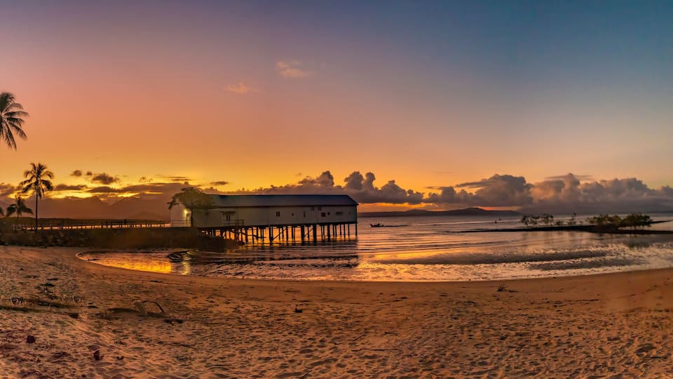 A beach with a wooden structure on stilts, palm trees, and a sunset over the water.