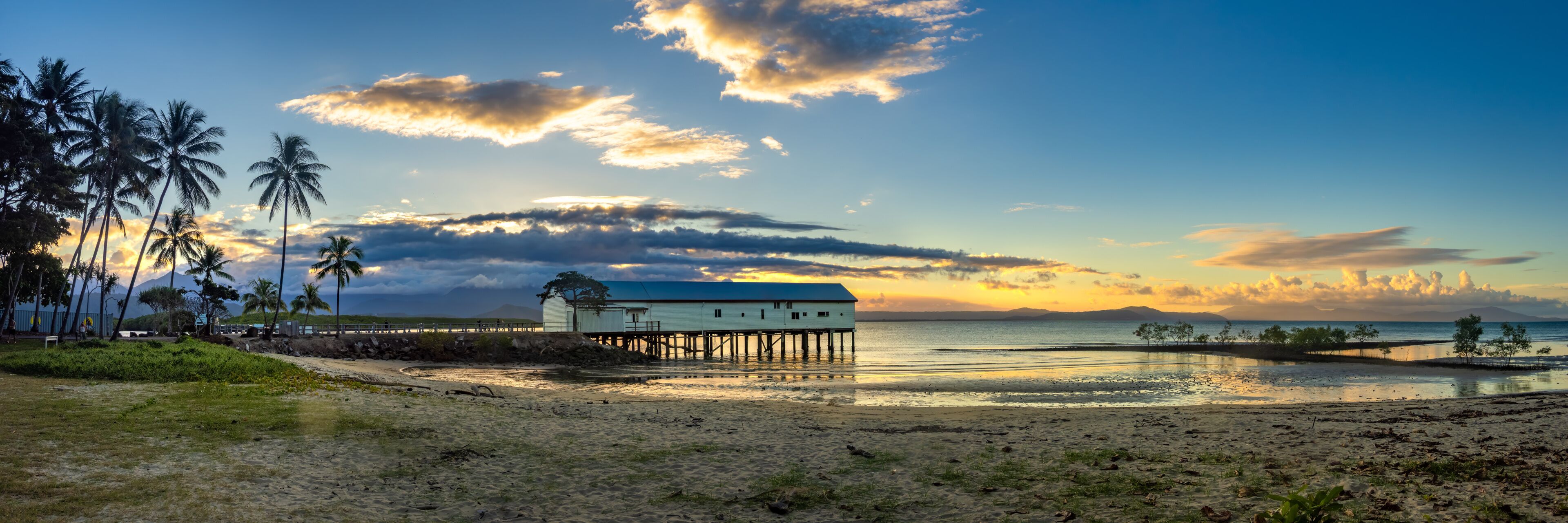 A beach with a wooden pier, palm trees, and a white building with a blue roof.