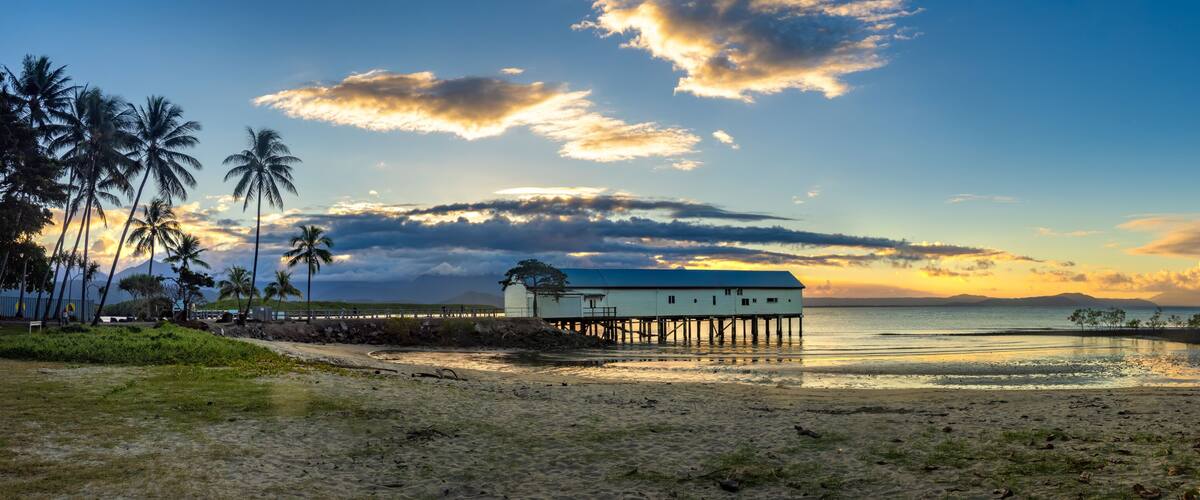 A beach with a wooden pier, palm trees, and a white building with a blue roof.