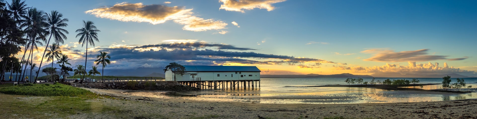 A beach with a wooden pier, palm trees, and a white building with a blue roof.