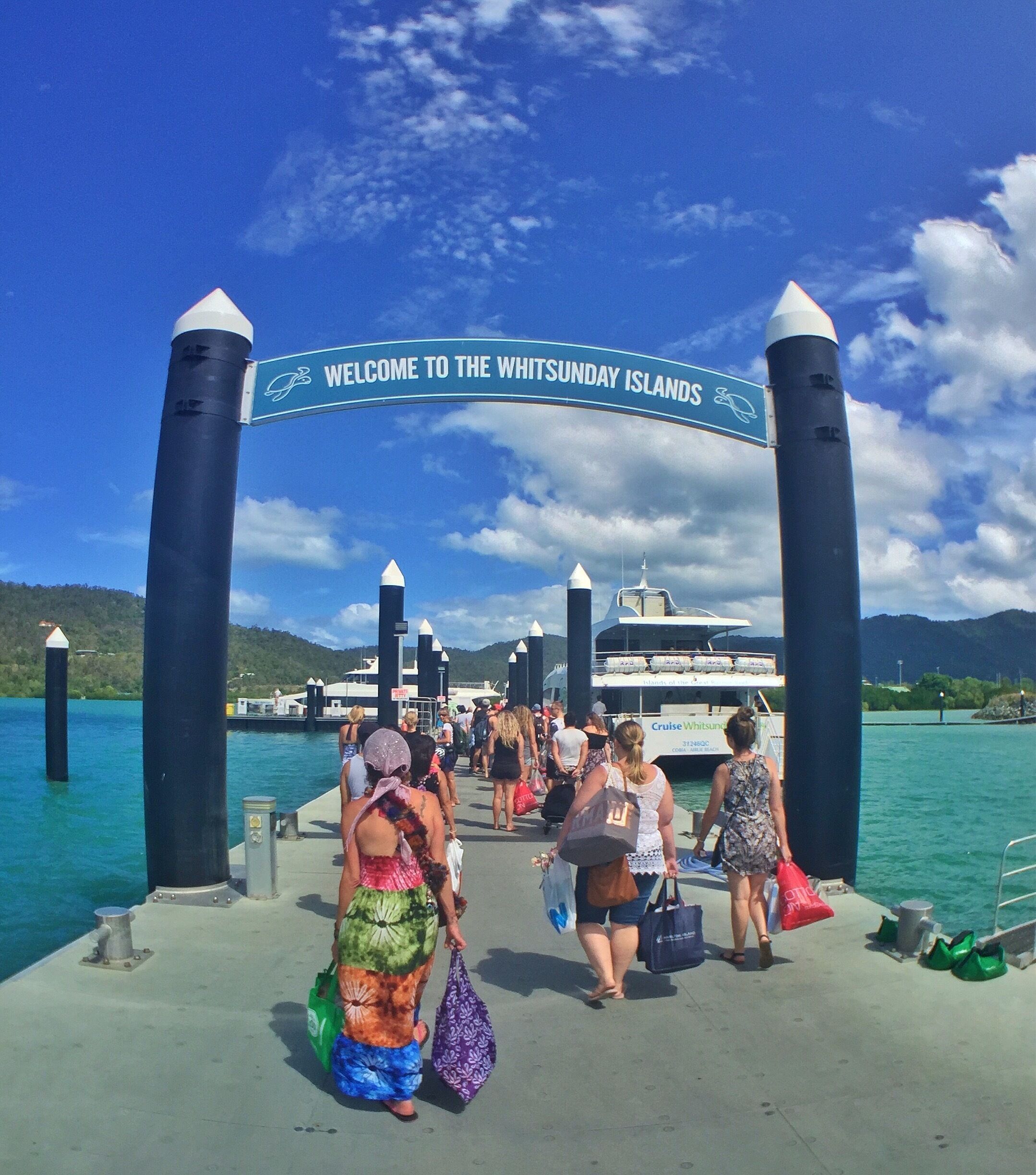 A pier with a 'Welcome to the Whitsunday Islands' sign, people walking, and a boat in the background.