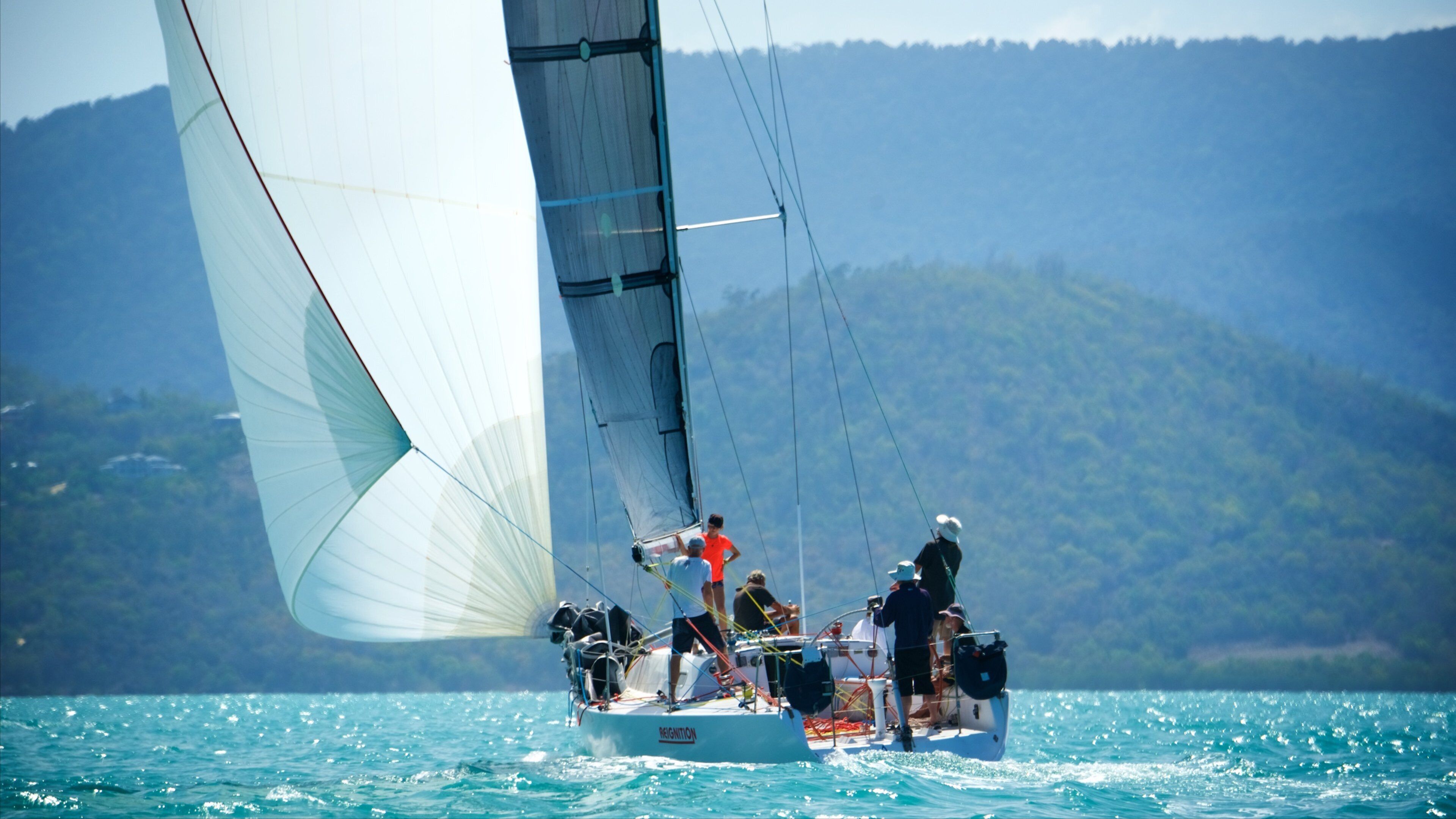 A sailboat with a crew on a calm sea with a mountainous backdrop.