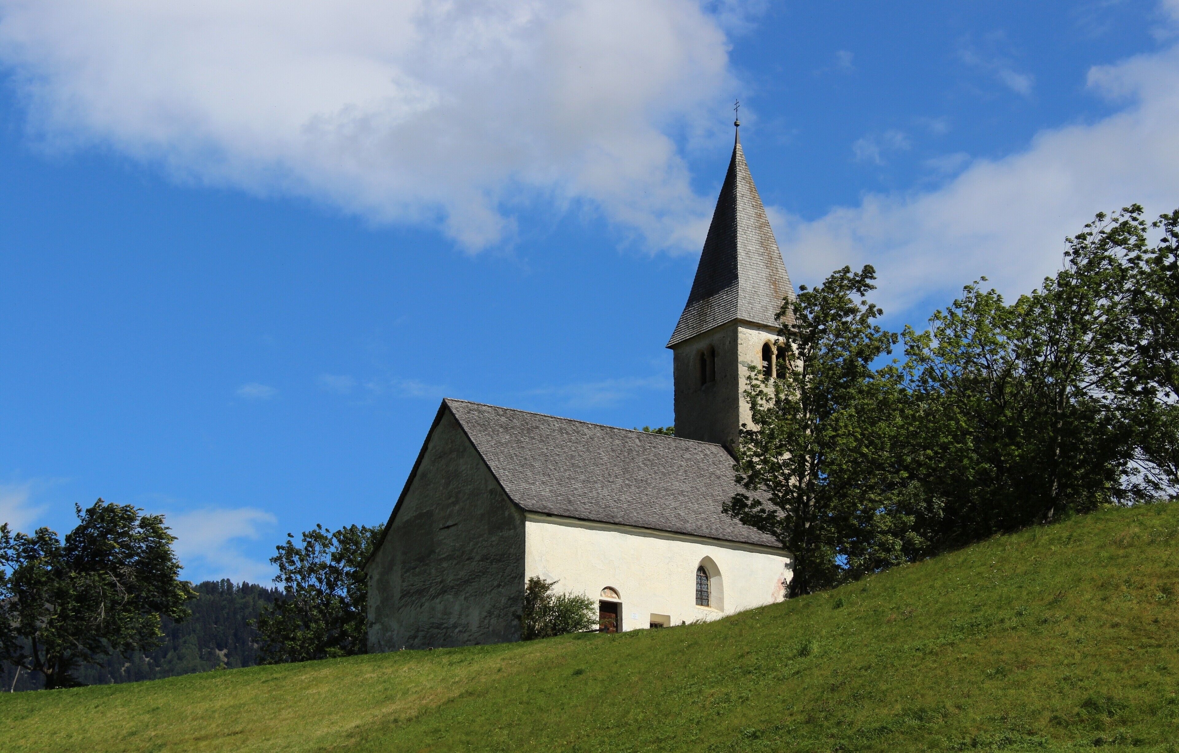 A church with a tall steeple on a grassy hill under a blue sky.