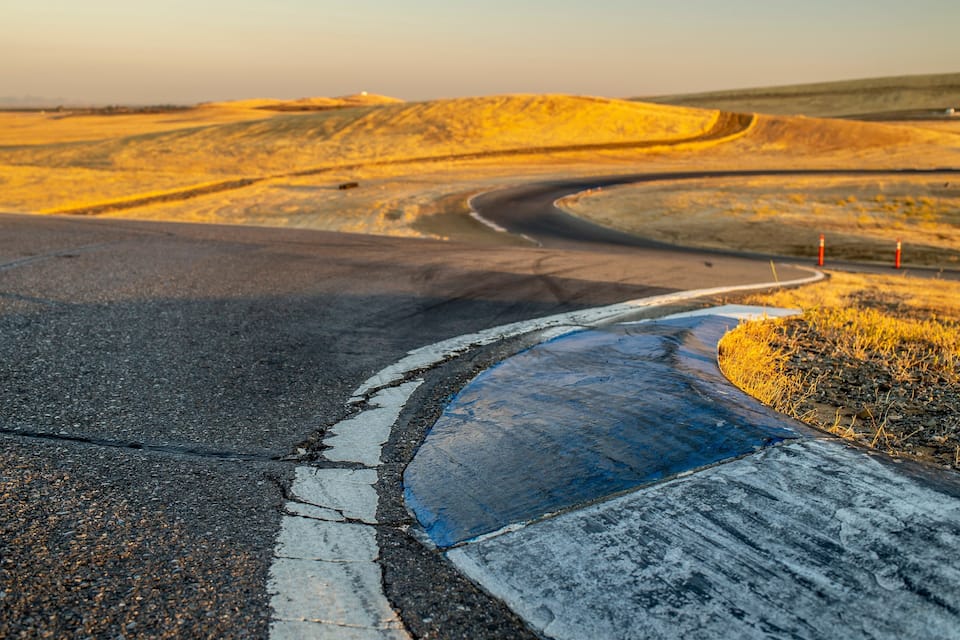 A winding road with a blue painted edge, surrounded by rolling hills and a sunset sky.