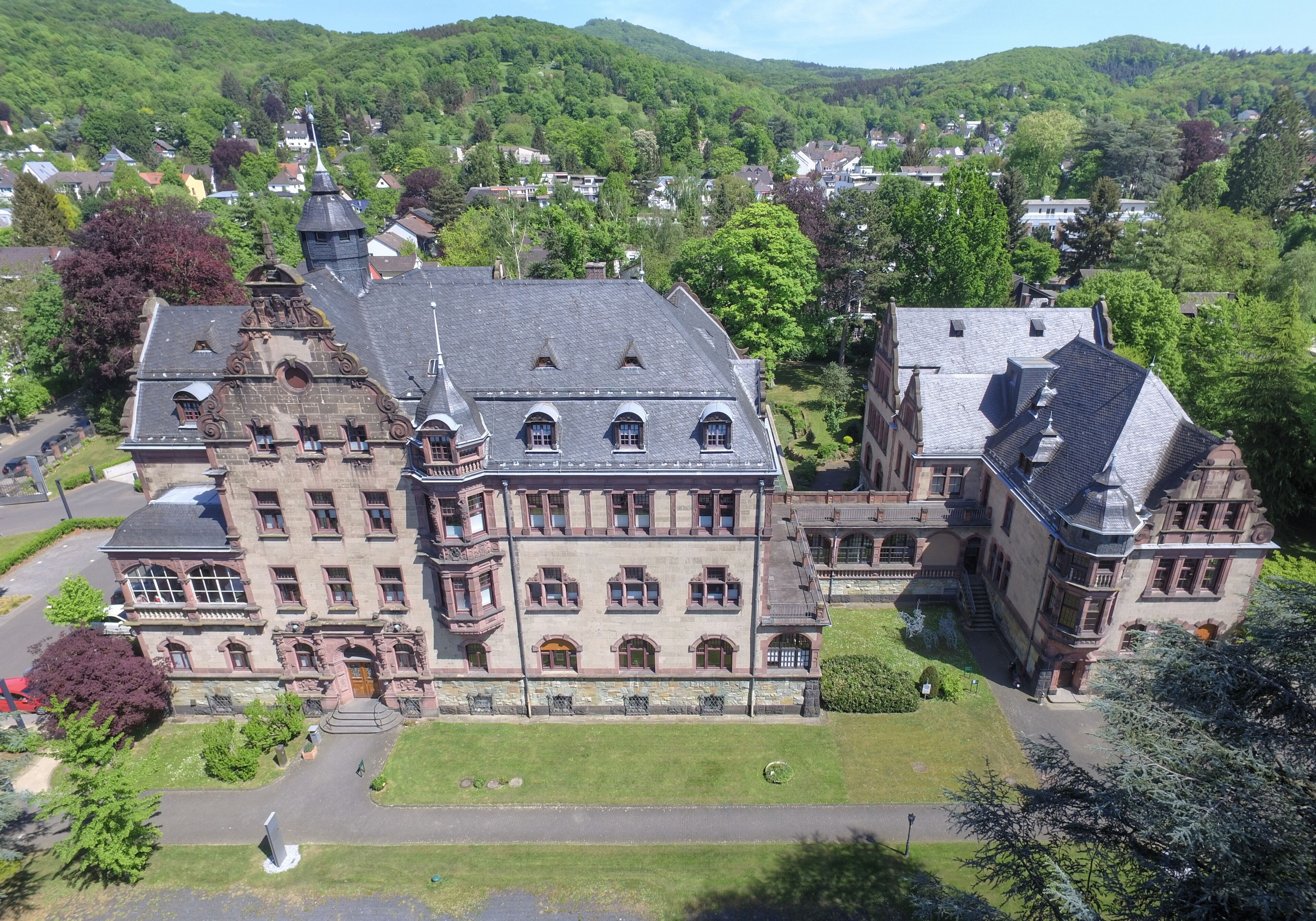 A large, historic building with a prominent tower and multiple windows, surrounded by lush greenery and hills.