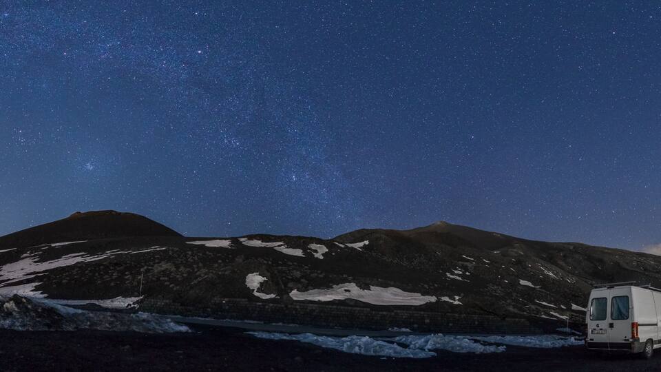 A white van parked in a snowy landscape with mountains under a starry night sky.