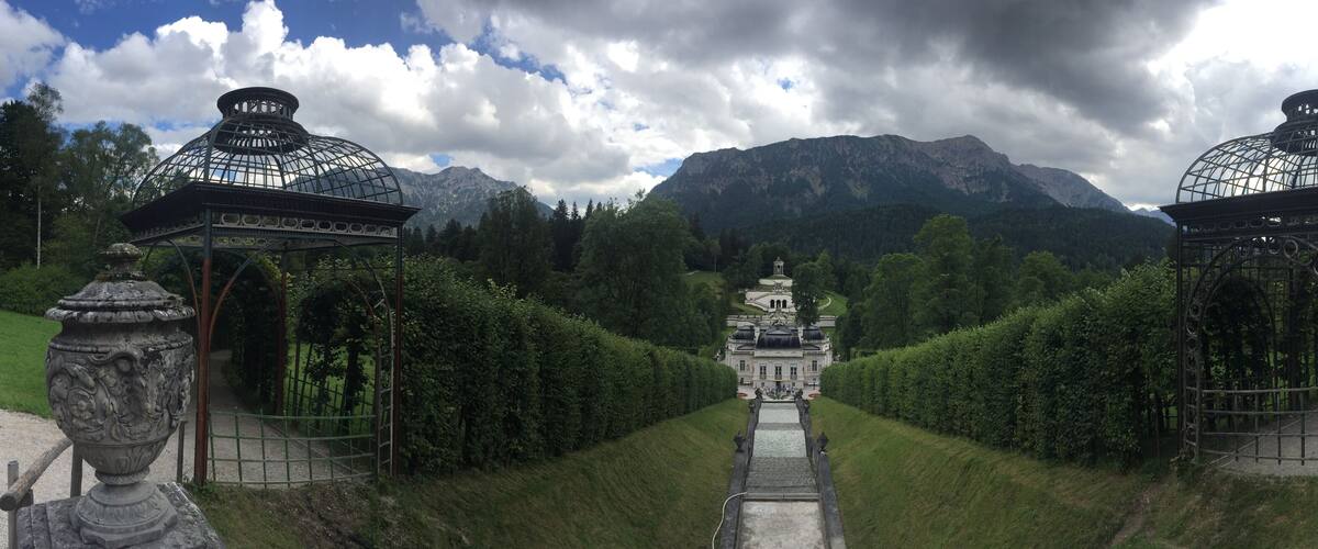 A pathway flanked by ornate lanterns leading to a central structure with a dome, surrounded by lush greenery and mountains in the background.