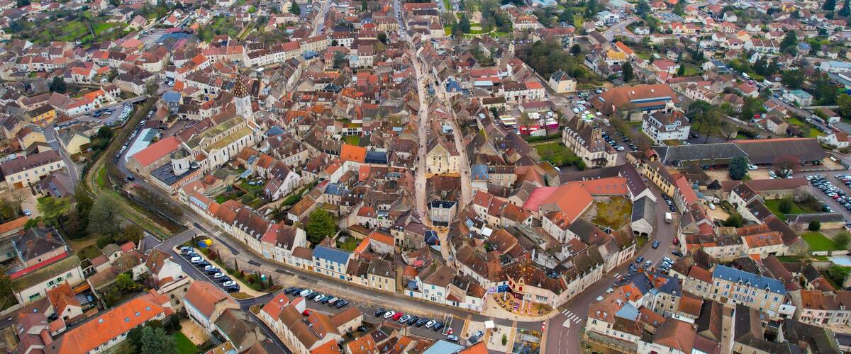 An aerial view of a densely packed town with red-roofed buildings, a central road, and surrounding fields.