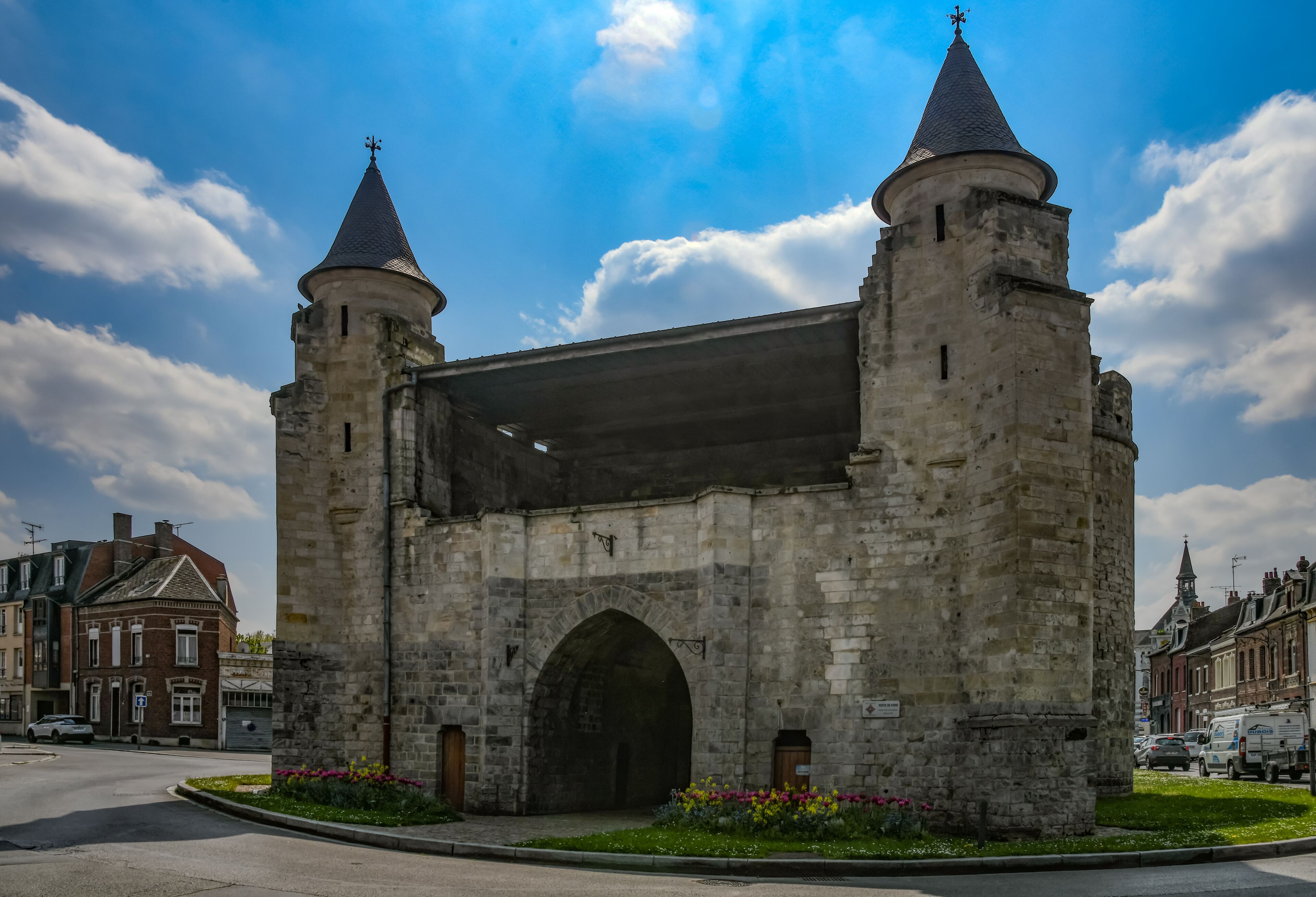 A stone archway with two towers, surrounded by a street and buildings.