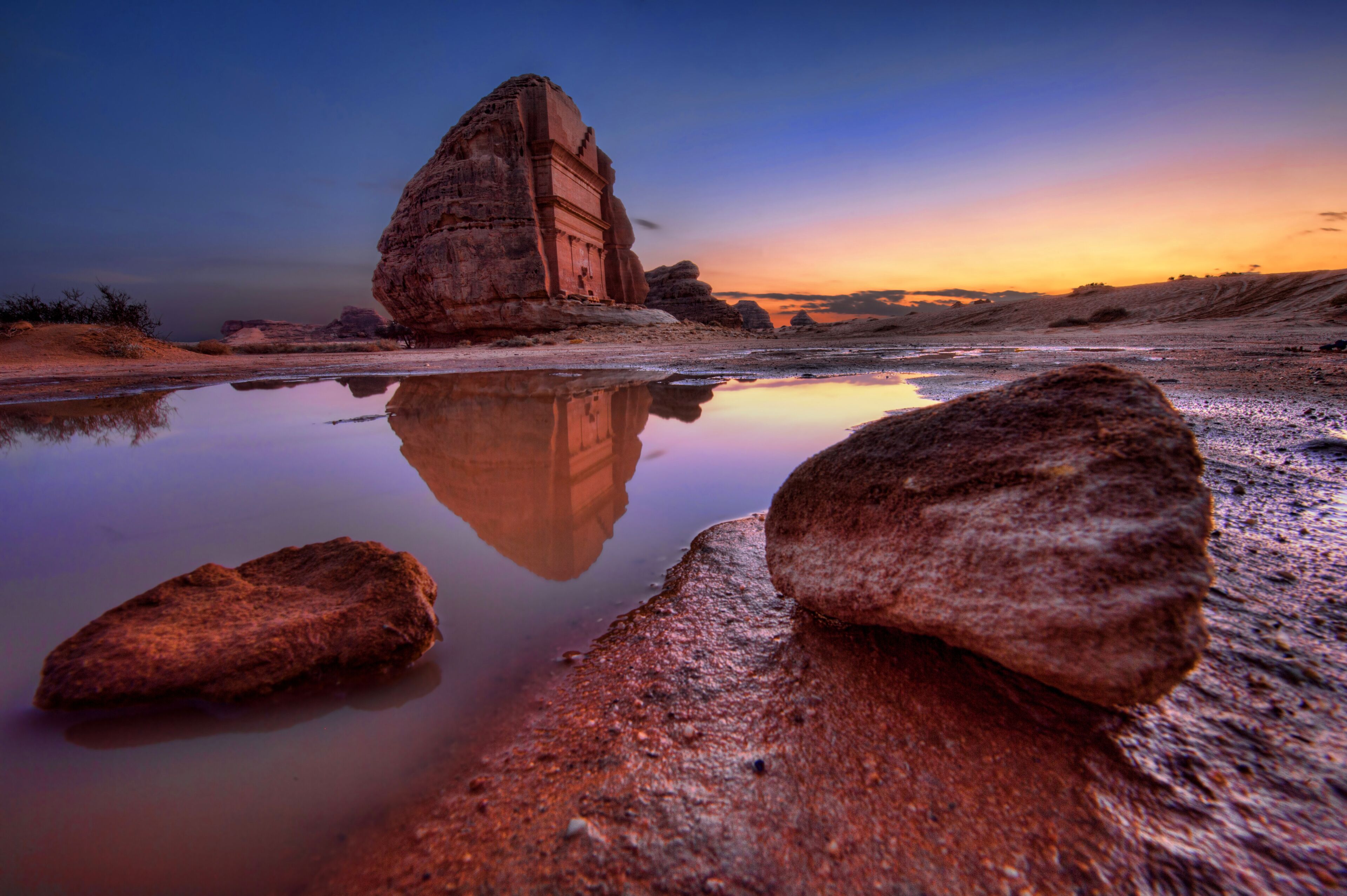 Formação rochosa refletida em águas rasas, com o céu ao pôr do sol como pano de fundo.