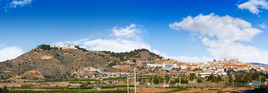 Una colina con un edificio en la cima, un pueblo en el valle y un cielo azul despejado.