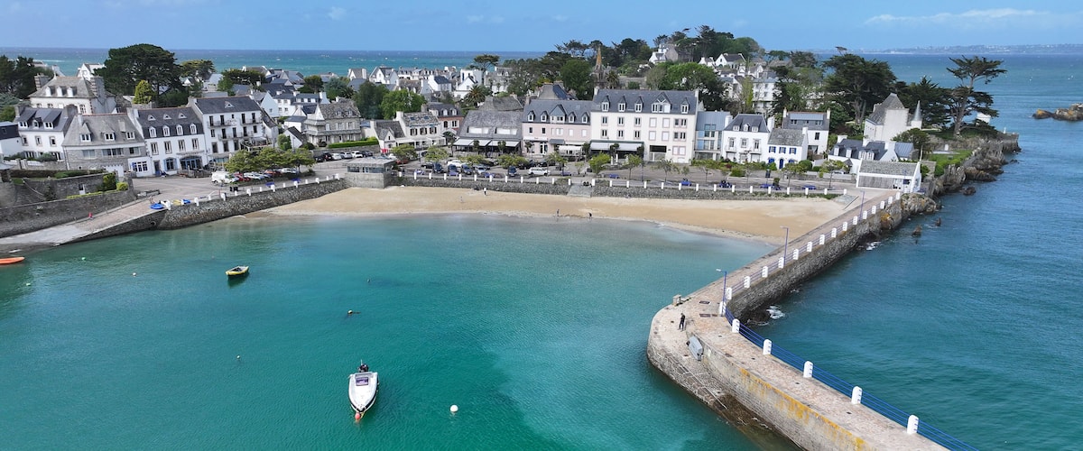 Aerial view of the picturesque harbor of Locquirec featuring a stone pier, sandy beach, and traditional coastal houses along the turquoise sea, Locquirec, Brittany, France.
