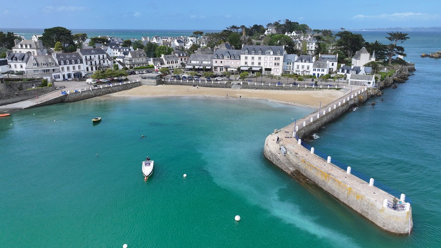 Aerial view of the picturesque harbor of Locquirec featuring a stone pier, sandy beach, and traditional coastal houses along the turquoise sea, Locquirec, Brittany, France.