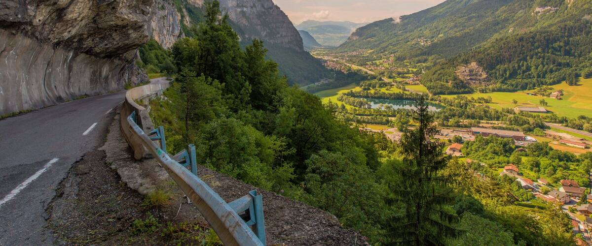 A winding mountain road with guardrails, surrounded by lush greenery and a valley with buildings and a lake.