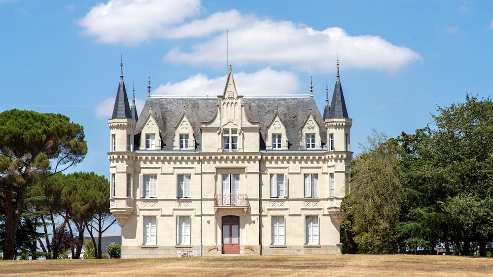 Un grand bâtiment historique doté de nombreuses tours et d’une flèche centrale, entouré d’arbres et d’un champ verdoyant.