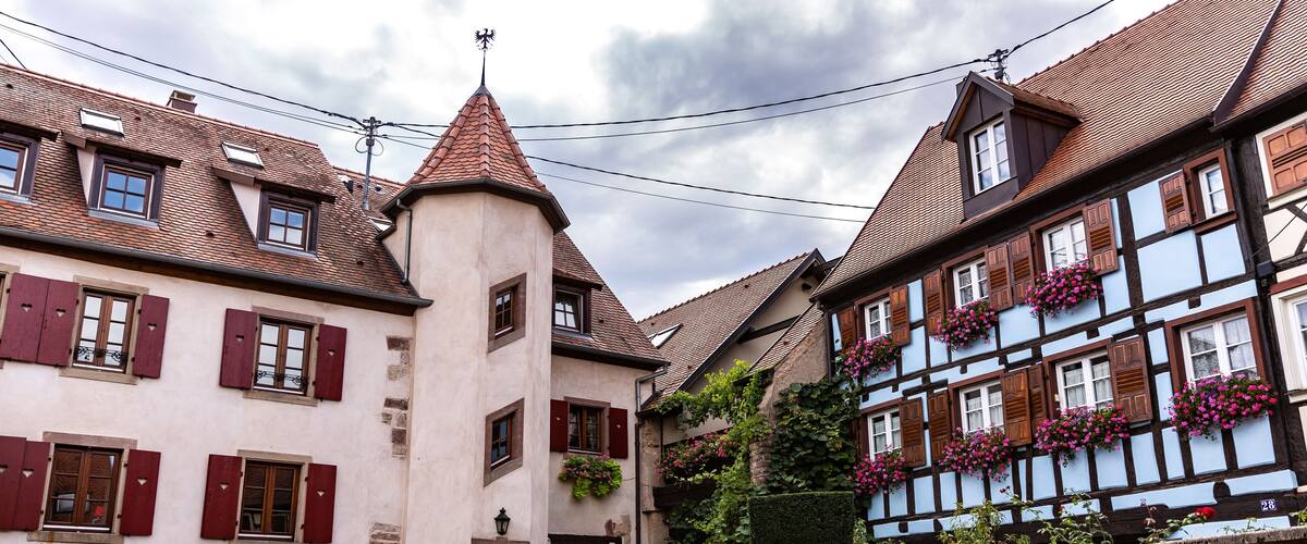A narrow cobblestone street flanked by traditional half-timbered houses with red shutters and a tower with a weather vane.