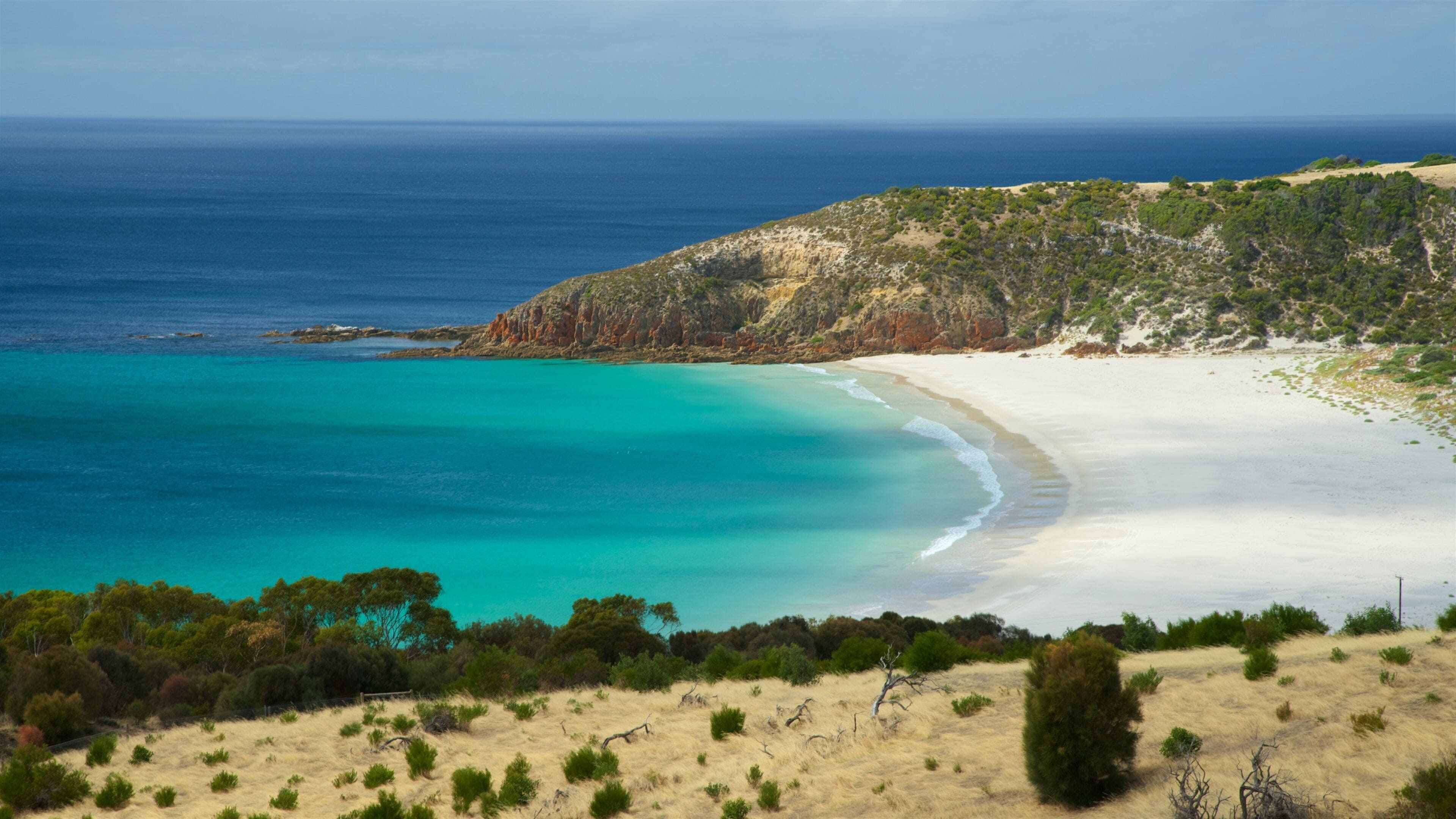 A coastal landscape with a sandy beach, turquoise waters, and green hills.