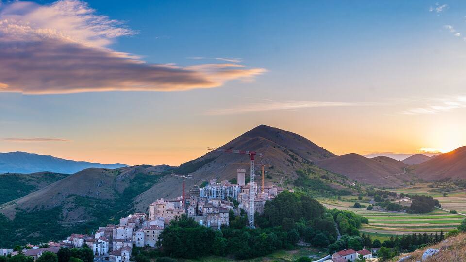 A mountainous landscape with a village at the base, a prominent peak, and a sunset in the background.