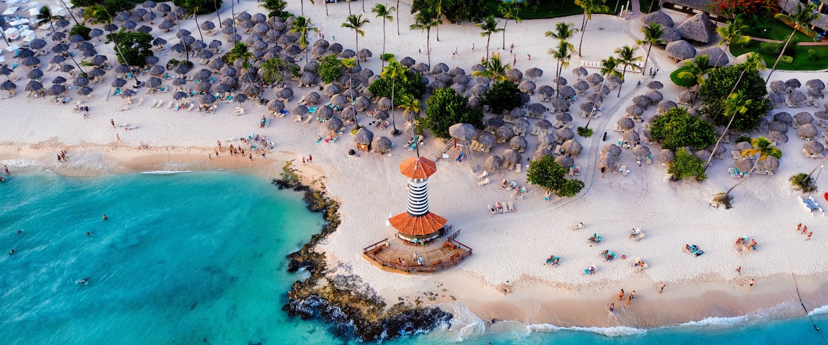 An aerial view of a beach with thatched umbrellas, palm trees, and a lighthouse.