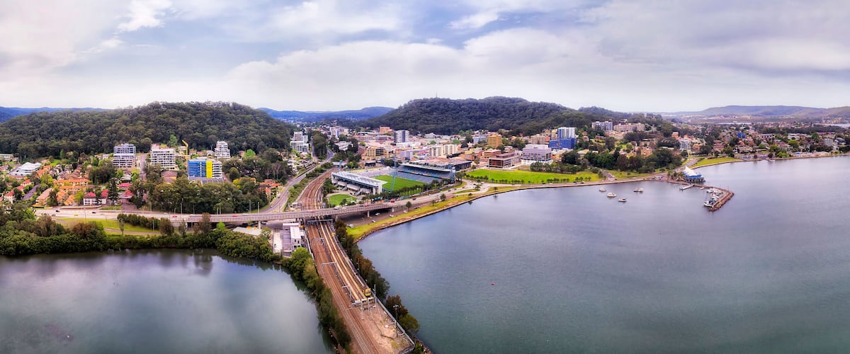 A riverside cityscape with a bridge, buildings, and a forested hill.